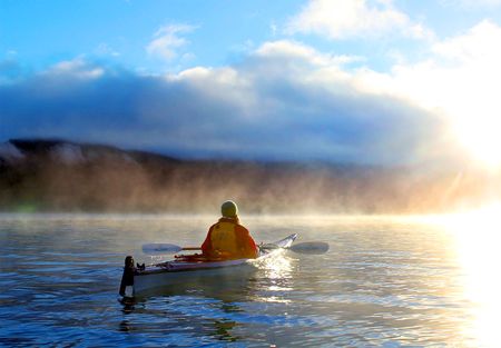 A person kayaks on calm waters under a misty sunrise, with clouds hovering low on the horizon.