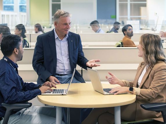 
                                                Three professionals, two with laptops, are conversing at a round table in a bright, modern office space.