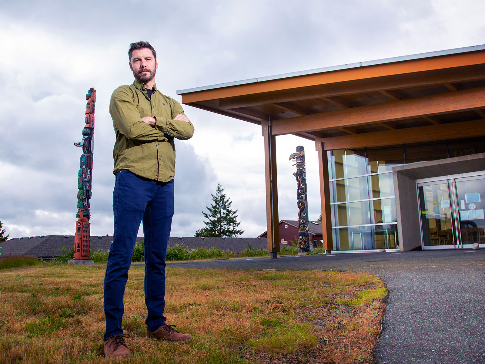 
                                A confident man with crossed arms stands in front of a modern building with large windows and two totem poles.