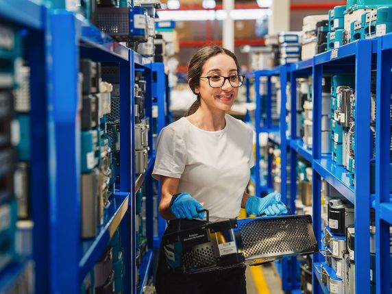 
                                                A woman wearing glasses, a white shirt, and blue gloves is smiling while holding a black basket in a warehouse with blue storage racks filled with electronic components.