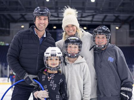 A happy family of five is posing together at an ice rink; the parents are smiling behind three children who are wearing hockey helmets, and the father is also wearing a helmet, indicating they are enjoying a day of ice hockey.