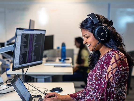 
                                                    A smiling woman wearing a floral dress and headphones is working on a laptop, with a dual monitor setup displaying code in the background, indicative of a software development or data analysis environment.