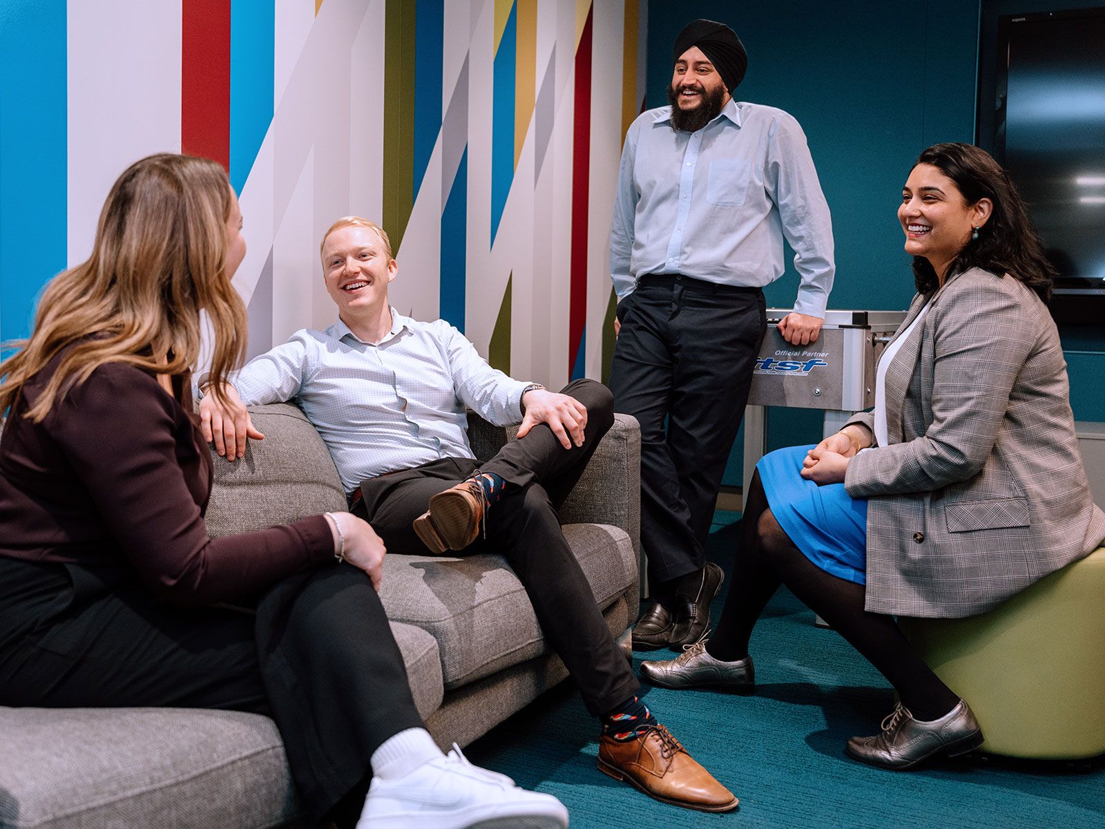 Four professionals are having a casual and cheerful conversation in a modern office lounge with colorful wall designs, with two seated on a sofa and one on an ottoman while the fourth stands, suggesting a relaxed and collaborative work environment.