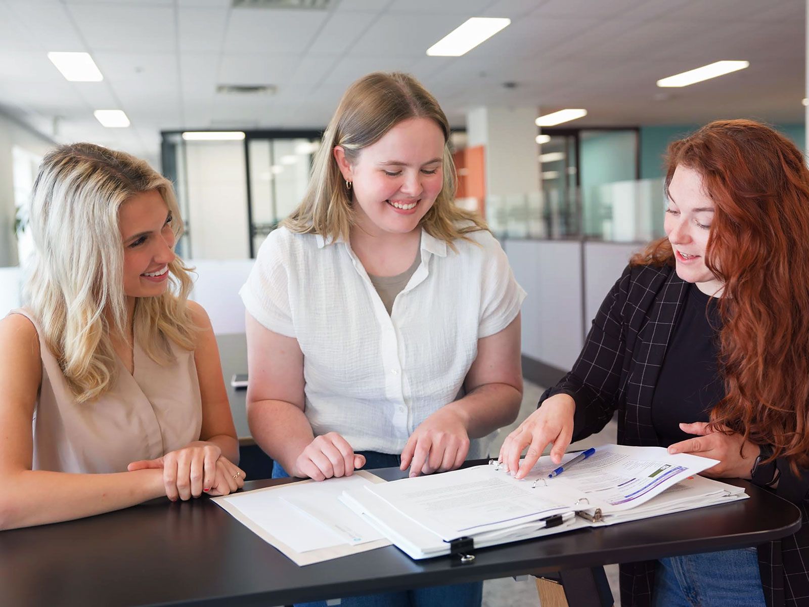 Three women smiling and collaborating over documents on a table in an office environment.
