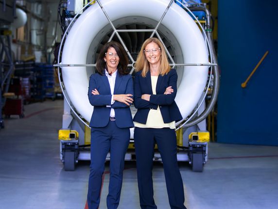 Two smiling women in business attire standing in front of a large industrial turbine engine in a manufacturing facility.