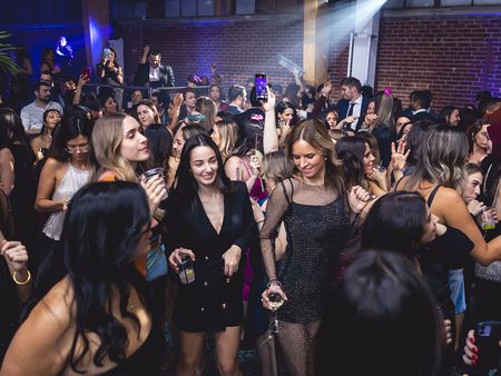 A lively party scene with a group of women dressed in evening wear, dancing and enjoying themselves in a crowded venue with brick walls and blue lighting.